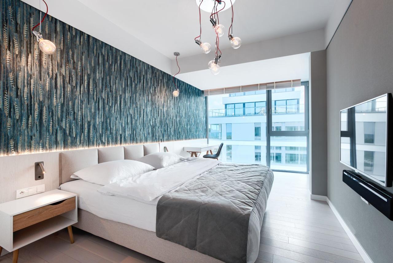 Nowoczesna sypialnia z tapetą w odcieniach niebieskiego, łóżkiem z szarą narzutą i białymi poduszkami, wiszącymi lampami z żarówkami Edisona, biurkiem przy oknie z widokiem na budynek...