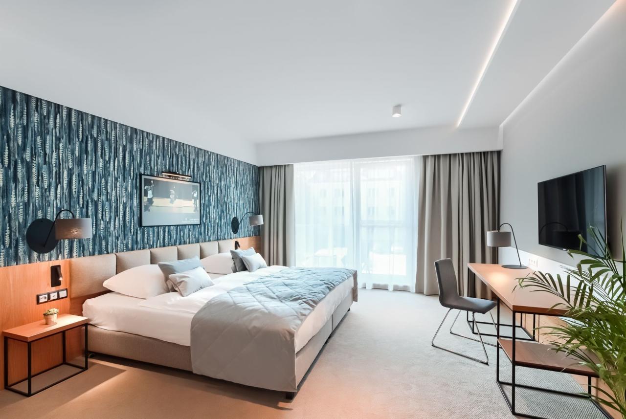 Nowoczesna sypialnia w hotelu z łóżkiem typu king-size, niebieską tapetą w motywy roślinne, biurkiem z lampką i telewizorem na ścianie.