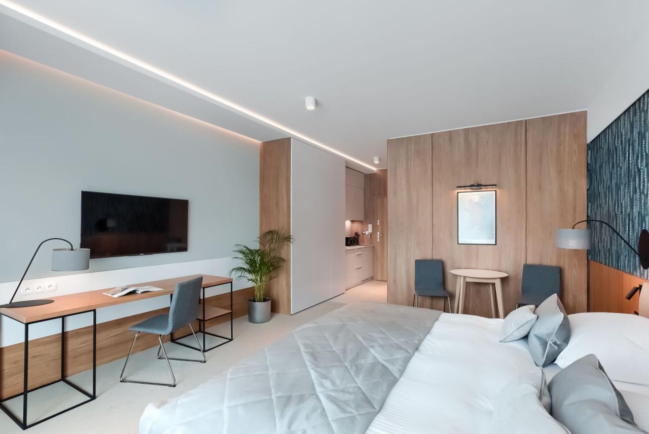 Nowoczesne studio z aneksem kuchennym, biurkiem do pracy, łóżkiem z szarą narzutą i dekoracyjną ścianą z niebieską tapetą.