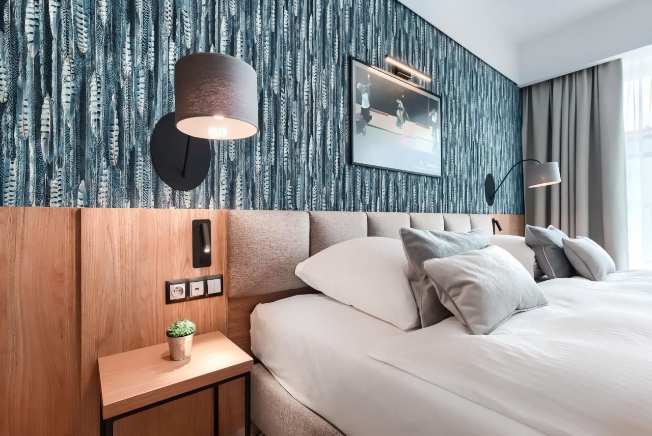 Nowoczesna sypialnia z łóżkiem kontynentalnym, zagłówkiem z paneli, tapetą w motywy piór, lampką nocną z abażurem i stolikiem nocnym z rośliną.