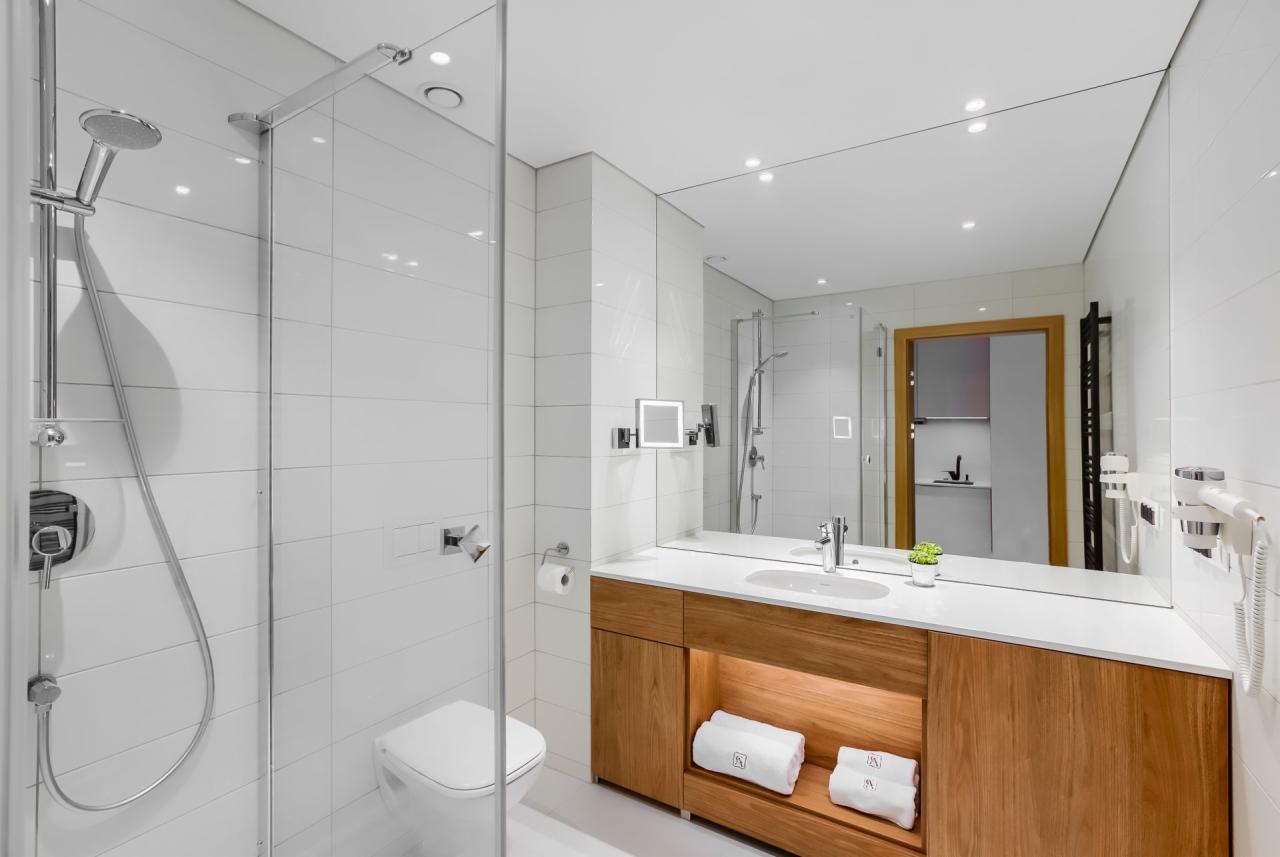 Nowoczesna łazienka z kabiną prysznicową, umywalką w drewnianej szafce podświetlanej od spodu i dużym lustrem w Koszalinie. Na blacie mała roślina doniczkowa, a pod umywalką ułożone białe ręczniki.