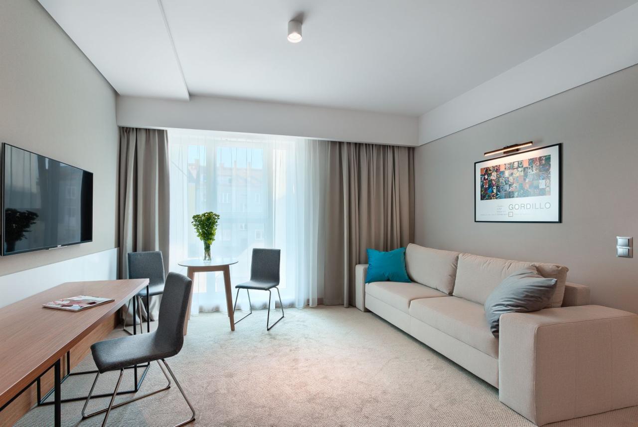 Jasne wnętrze pokoju hotelowego z beżową sofą, drewnianym biurkiem, okrągłym stolikiem z bukietem kwiatów i plakatem na ścianie.