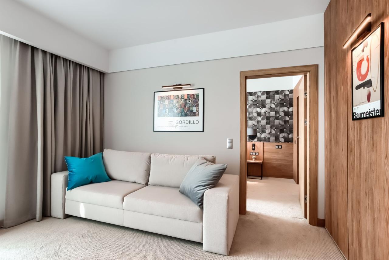 Jasny salon z rozkładaną sofą, turkusową i szarą poduszką, plakatami na ścianie i widokiem na sypialnię z geometryczną tapetą.