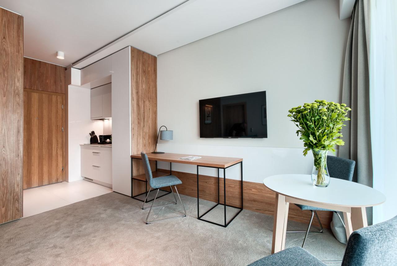 Nowoczesne wnętrze apartamentu z aneksem kuchennym, biurkiem z lampką, okrągłym stołem z bukietem zielonych kwiatów i telewizorem na ścianie.