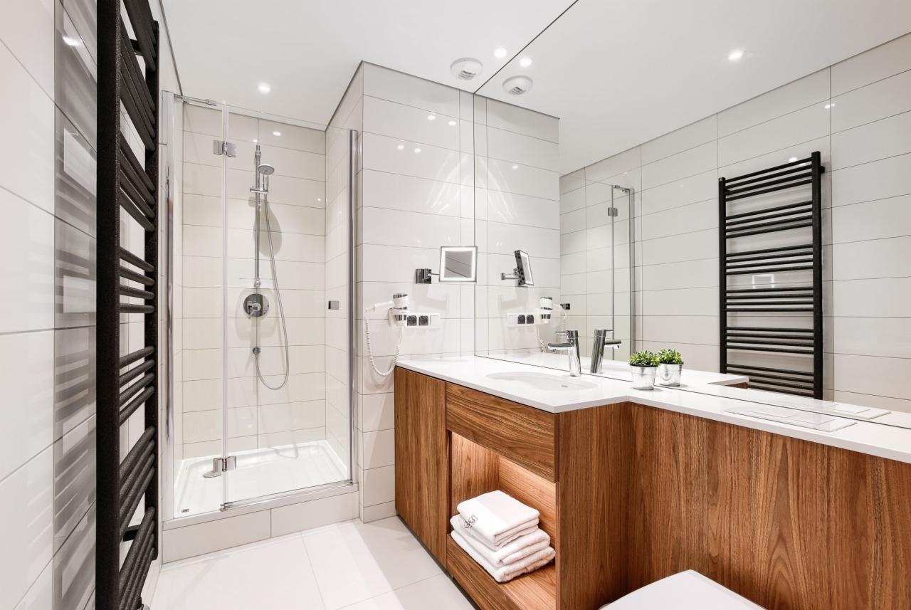 Nowoczesna łazienka z kabiną prysznicową, grzejnikiem drabinkowym i szafką z drewnianym fornirem pod umywalką, oświetlona punktowo.