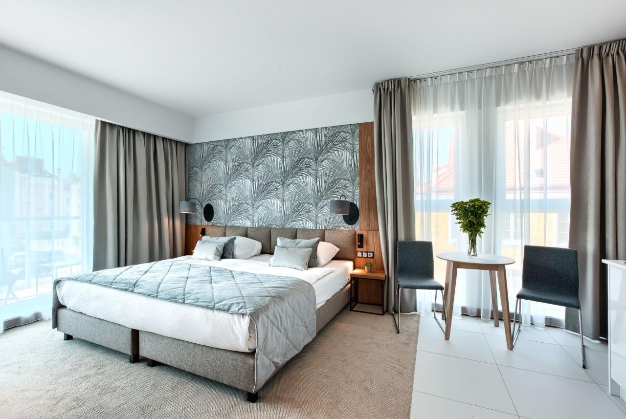 Nowoczesna sypialnia z łóżkiem typu king-size, tapetą w liście palmowe i stolikiem z kwiatami przy oknie.