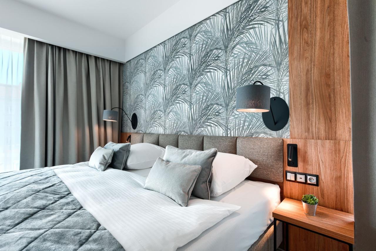 Nowoczesna sypialnia z tapetą w motywy roślinne, szarą pościelą i lampkami ściennymi z abażurami, akcentowana drewnianym panelem i małą rośliną doniczkową na stoliku nocnym.