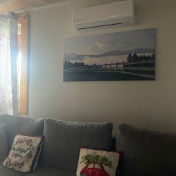 Światło Bracia Drewni - Biała klimatyzacja ścienna nad obrazem z krajobrazem w salonie z szarą sofą i świątecznymi poduszkami, drewniany sufit i okno z zasłonami w tle.