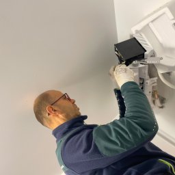 Światło Bracia Drewni - Instalator w okularach montuje klimatyzator na białym suficie. Widoczny biały moduł klimatyzacji i fragment instalacji. Ujęcie z dołu, podkreślające etap montażu.