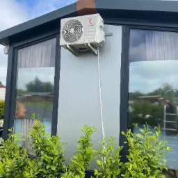 Biały klimatyzator Rotenso zamontowany na szarej ścianie budynku z dużymi oknami, widoczne zielone krzewy u podstawy ściany. Darłówko.