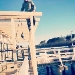 Konstrukcja drewniana w trakcie budowy nad wodą, dwóch robotników pracuje na niższym poziomie, trzeci stoi na górnej belce, w tle widoczny las i błękitne niebo.