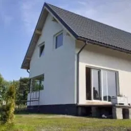 Nowoczesny, biały dom jednorodzinny z ciemną dach&oacute;wką, widoczny z perspektywy ogrodu, z osobą stojącą na tarasie i roślinami w donicach.