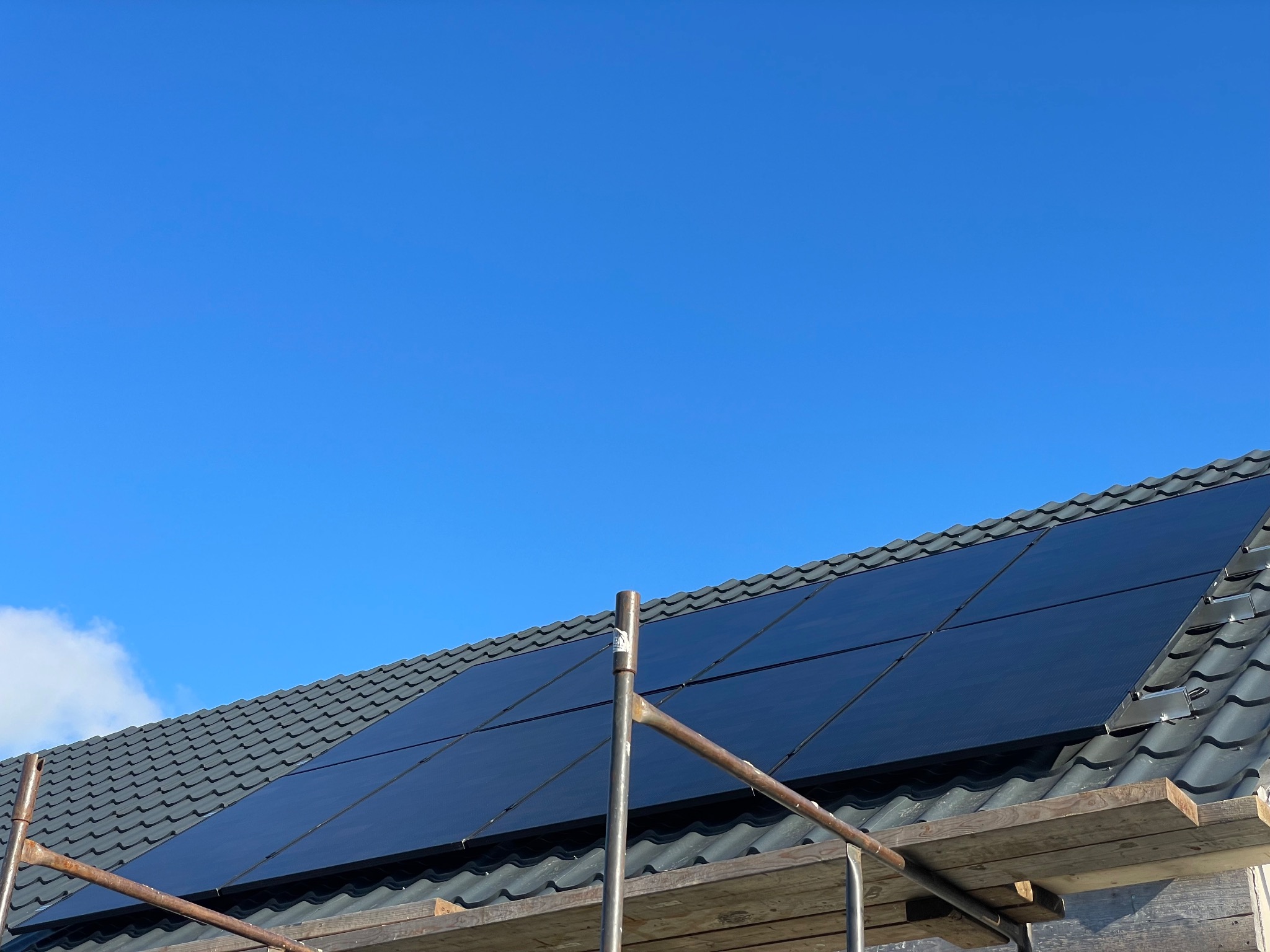 Montaż paneli fotowoltaicznych na dachu krytym szarą dachówką, widoczne rusztowanie i błękitne niebo.