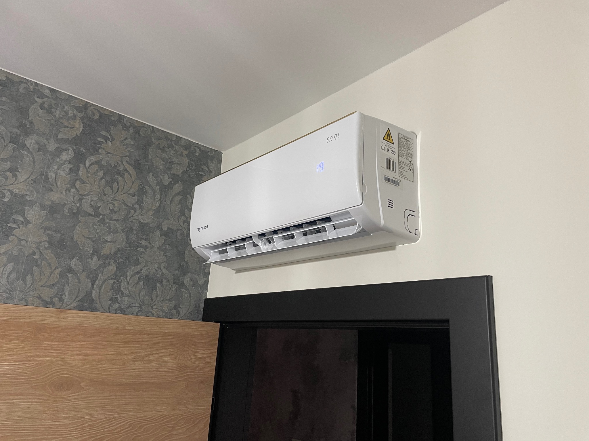 Biały klimatyzator marki Rotenso zamontowany na ścianie nad czarnymi drzwiami, widoczny fragment wzorzystej tapety i drewnianego elementu wykończenia.
