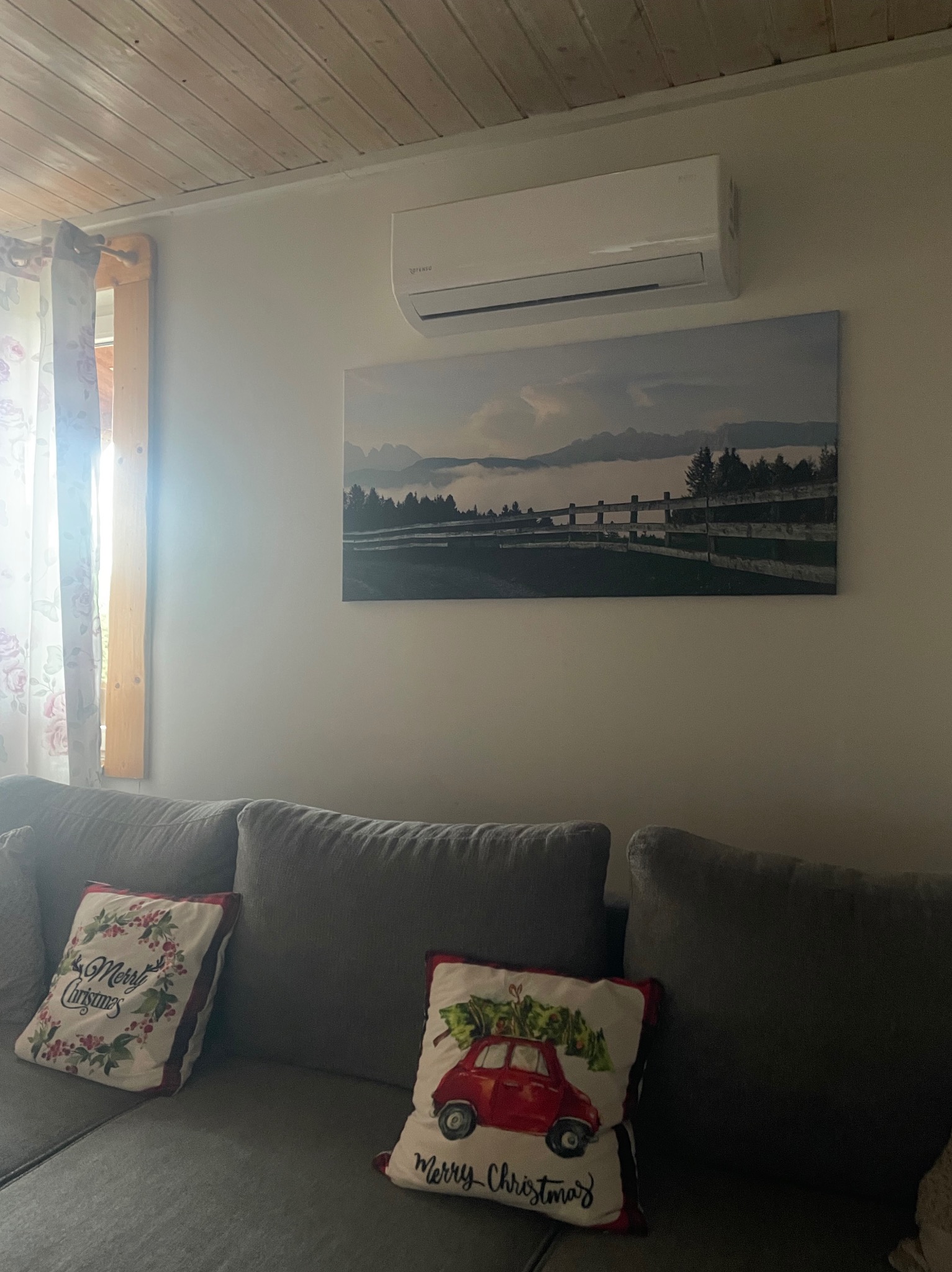 Biała klimatyzacja ścienna nad obrazem z krajobrazem w salonie z szarą sofą i świątecznymi poduszkami, drewniany sufit i okno z zasłonami w tle.