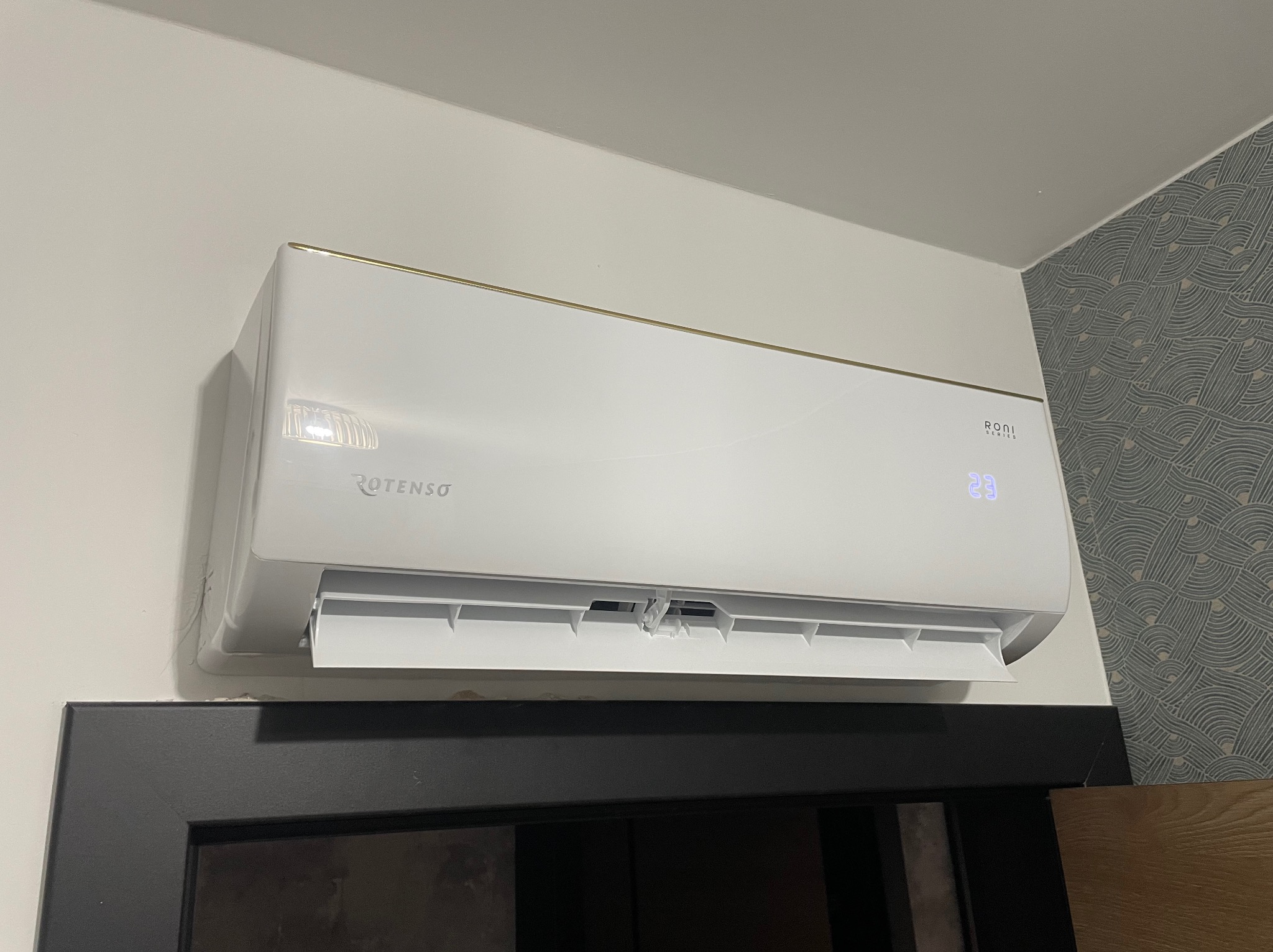 Biały klimatyzator Rotenso z delikatnym złotym paskiem, zamontowany na ścianie z wzorzystą tapetą, widoczny nad ciemną framugą drzwi. Wyświetlacz pokazuje 23 stopnie.