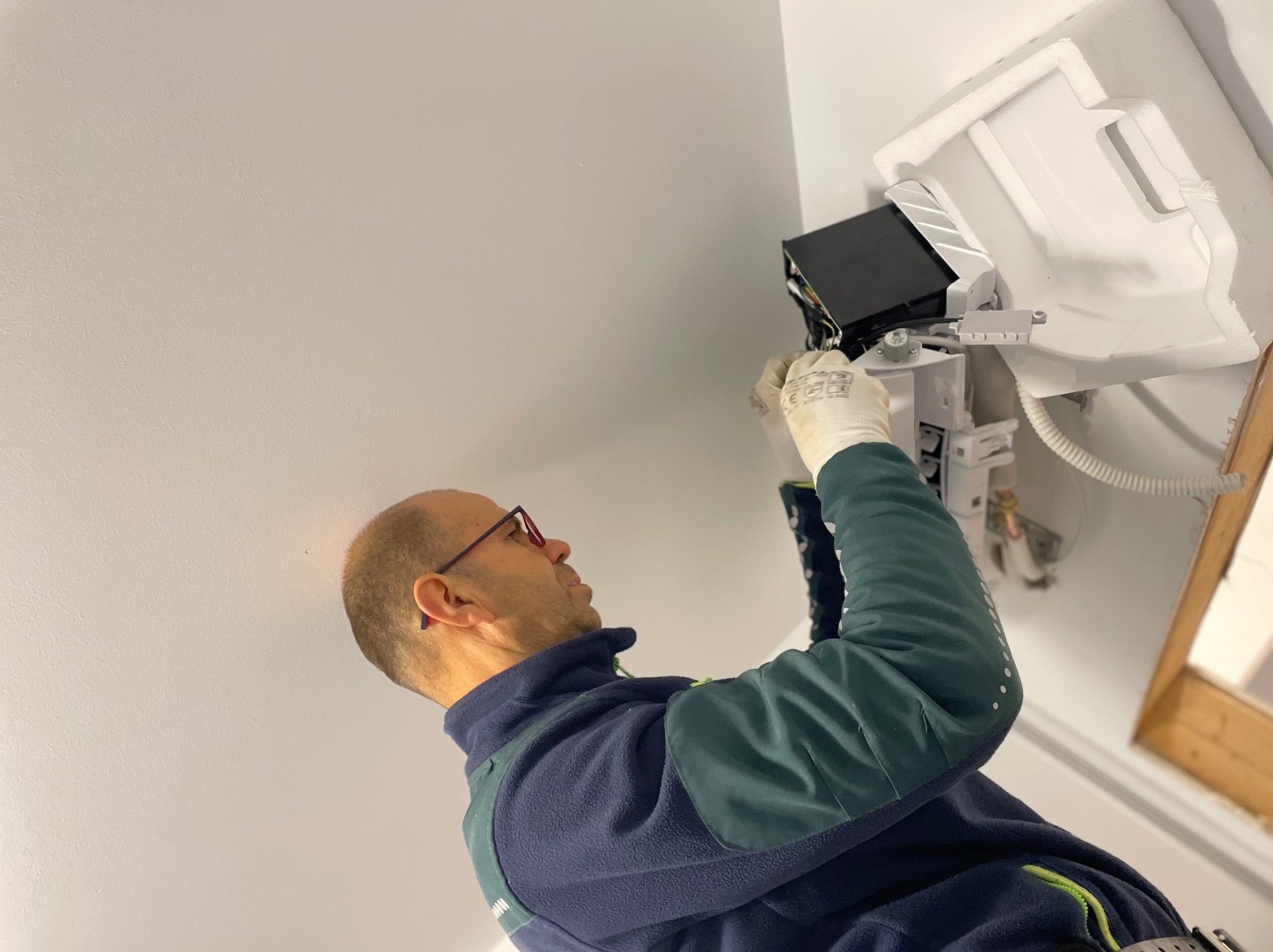 Instalator w okularach montuje klimatyzator na białym suficie. Widoczny biały moduł klimatyzacji i fragment instalacji. Ujęcie z dołu, podkreślające etap montażu.