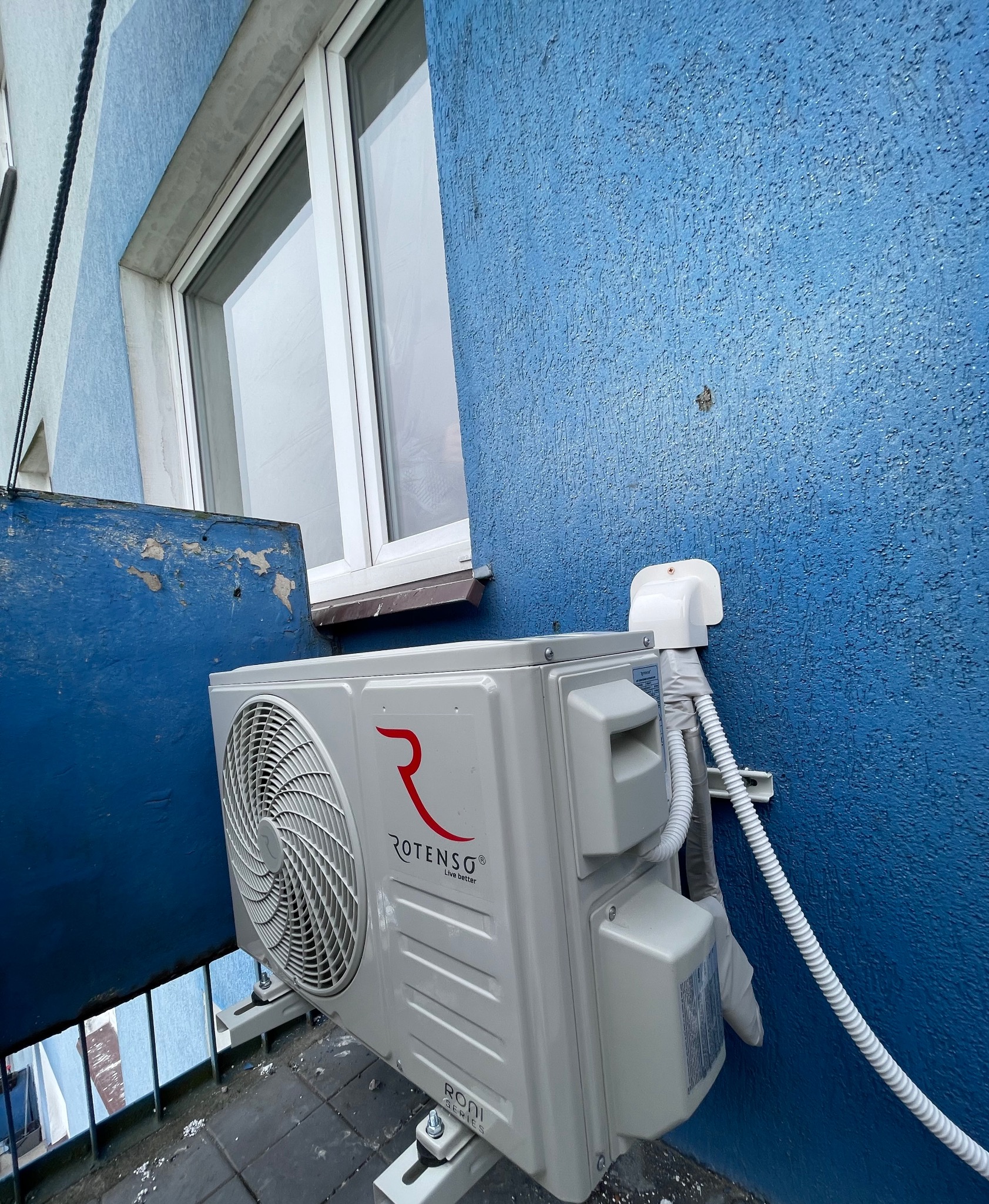 Jednostka zewnętrzna klimatyzacji Rotenso zamontowana na balkonie budynku wielomieszkaniowego z niebieską elewacją. Widoczny przewód i uchwyt montażowy.
