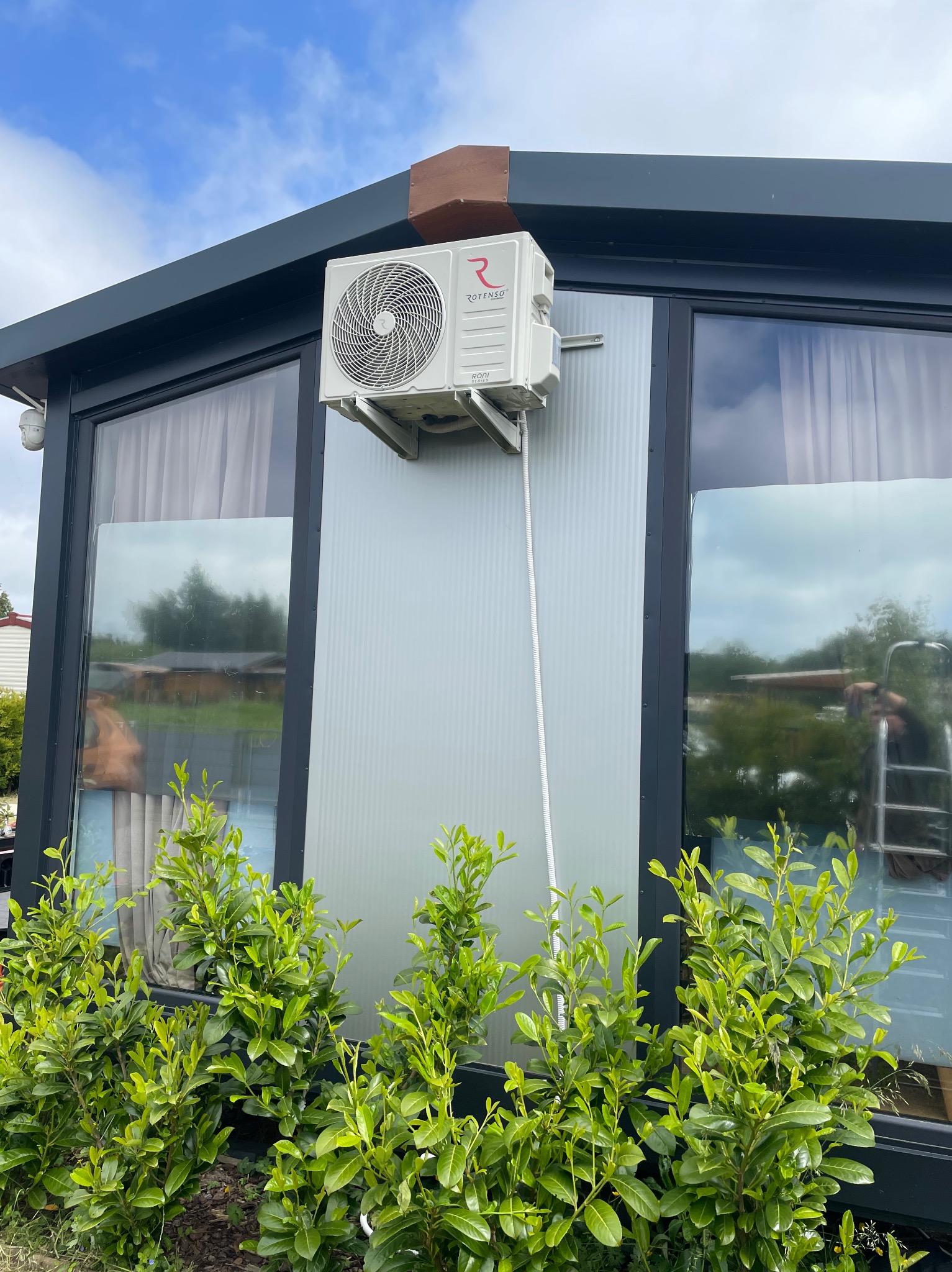 Biały klimatyzator Rotenso zamontowany na szarej ścianie budynku z dużymi oknami, widoczne zielone krzewy u podstawy ściany. Darłówko.