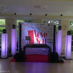 Scena z oświetleniem LED w fioletowych barwach, nagłośnienie i dekoracja z napisem 'Love You' w różnych językach, przygotowana na imprezę w jasnym wnętrzu.