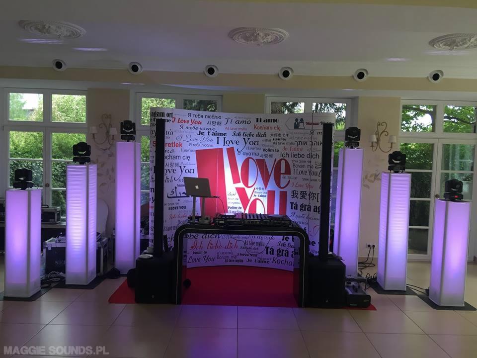 Scena z oświetleniem LED w fioletowych barwach, nagłośnienie i dekoracja z napisem 'Love You' w różnych językach, przygotowana na imprezę w jasnym wnętrzu.