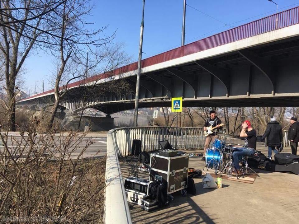 Zespół muzyczny grający pod mostem, perkusja w kolorze niebieskim, gitara elektryczna, nagłośnienie, znak drogowy w tle.