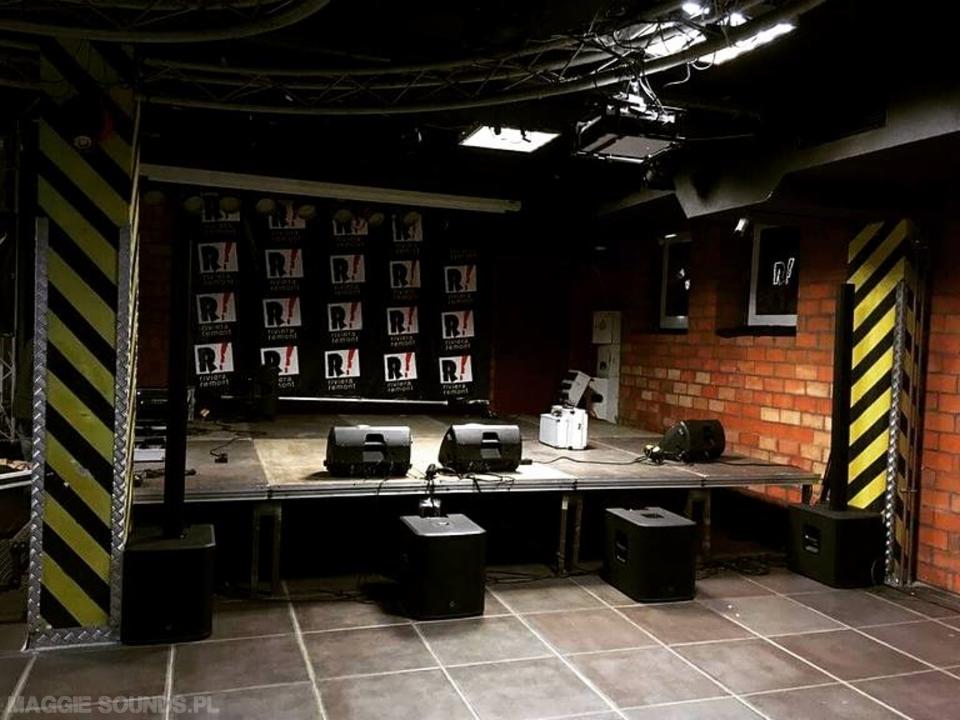 Scena z nagłośnieniem i oświetleniem w ciemnym pomieszczeniu z ceglaną ścianą i kolumnami w żółto-czarne pasy.