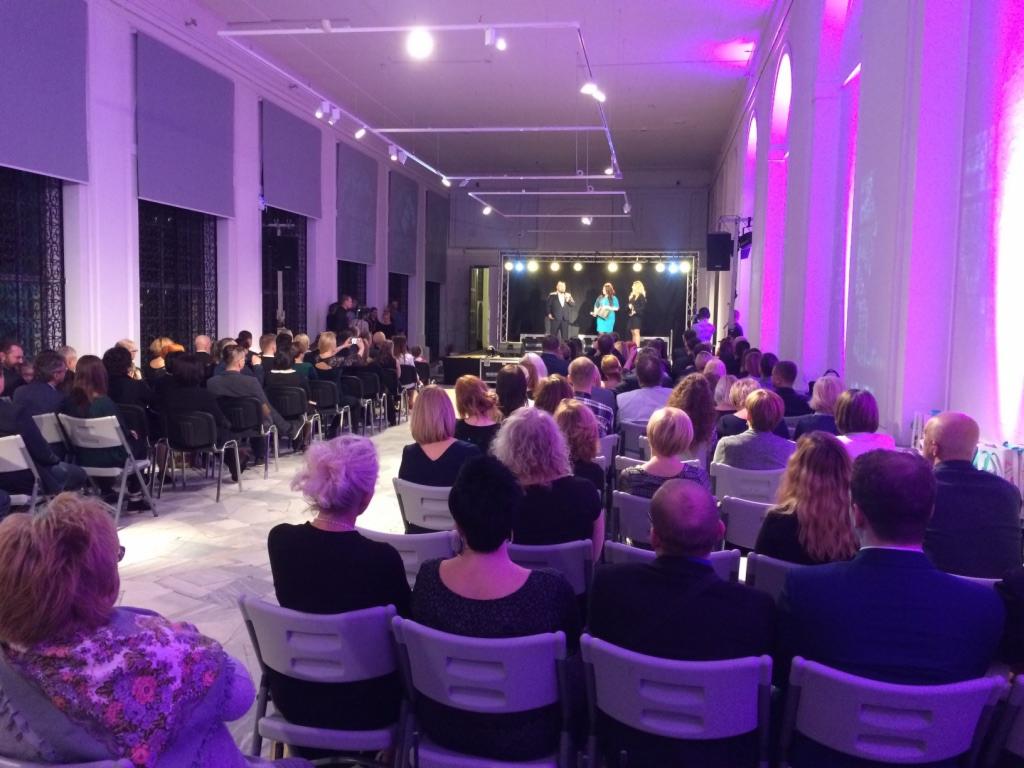 Widok sali konferencyjnej z publicznością siedzącą na krzesłach, scena z oświetleniem i występującymi osobami w tle, fioletowe oświetlenie architektoniczne sali.