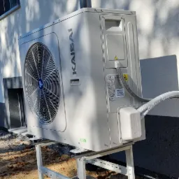 Zewnętrzna jednostka klimatyzacji Kaisai zamontowana na białym, metalowym stelażu przy ścianie budynku, z widocznym przewodem i cieniem drzewa na elewacji.