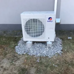 Zewnętrzna jednostka klimatyzacji Gree, umieszczona na betonowych blokach i otoczona szarym żwirem, na tle szarej ściany budynku z widocznym fragmentem rynny.