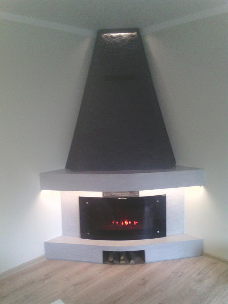 Narożny kominek z czarnym, stożkowym obudowaniem, podświetlony od góry i od dołu, z paleniskiem imitującym ogień, na tle jasnych ścian i drewnopodobnej podłogi.