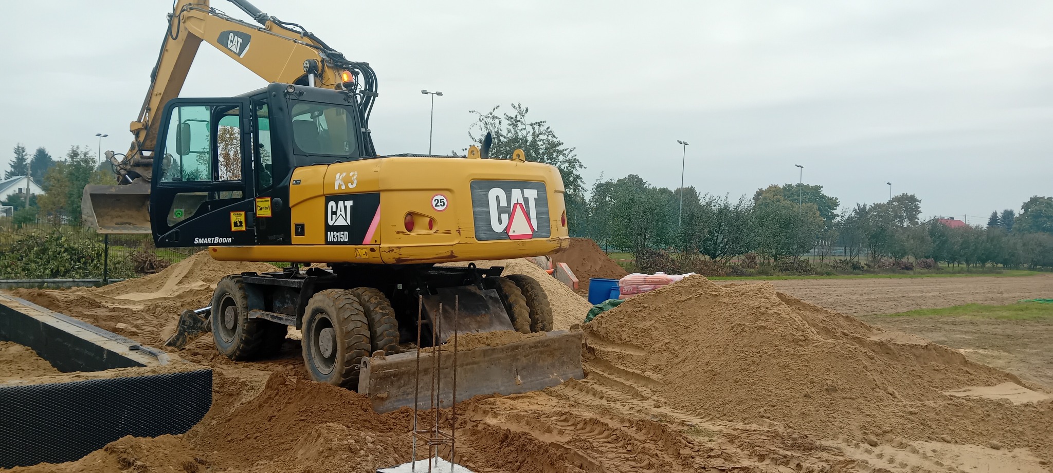 Żółta koparka kołowa CAT M315D na placu budowy, z widocznymi stertami piasku i elementami fundamentów. Maszyna gotowa do pracy przy niwelacji terenu.