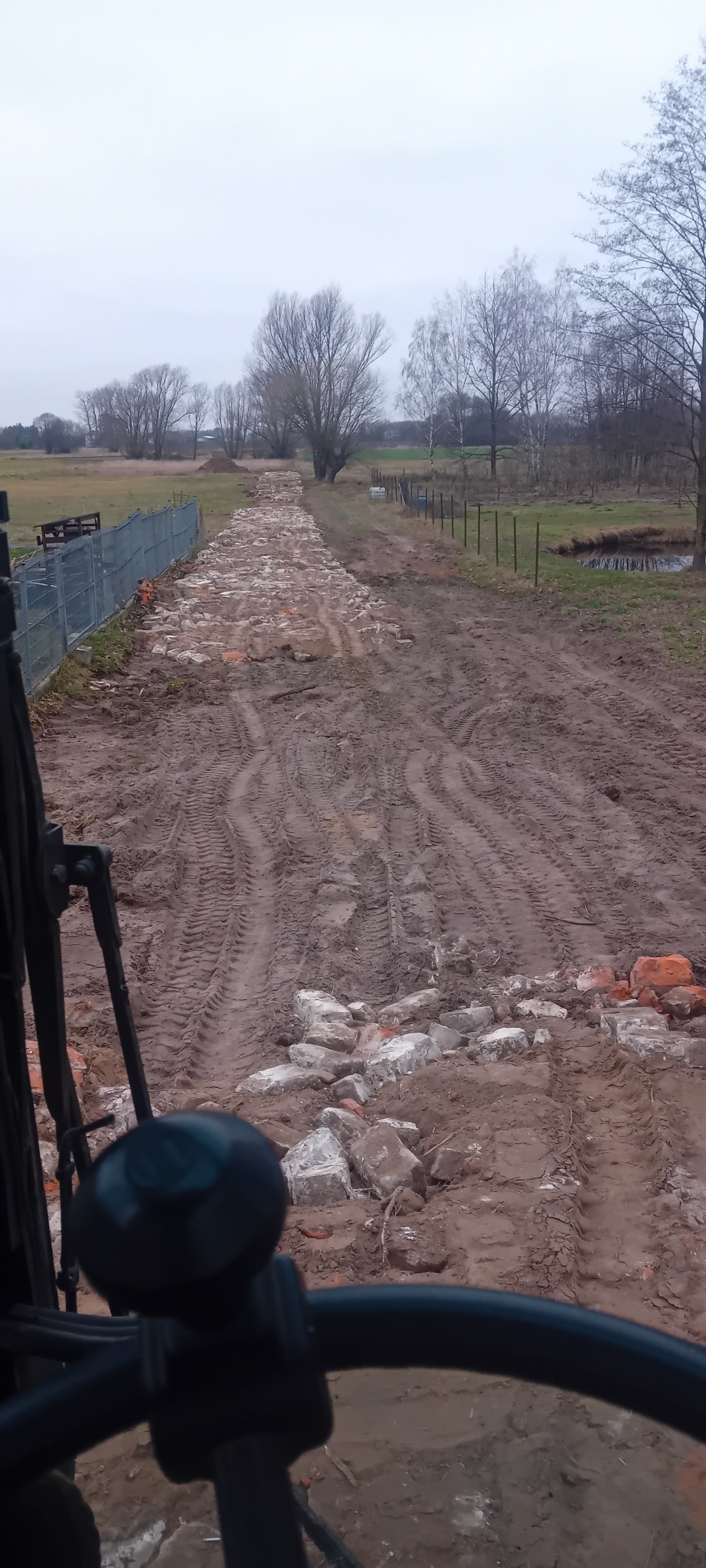 Widok z kabiny maszyny na błotnistą drogę z gruzem, prawdopodobnie w trakcie utwardzania. W tle drzewa i ogrodzenie, pochmurne niebo.