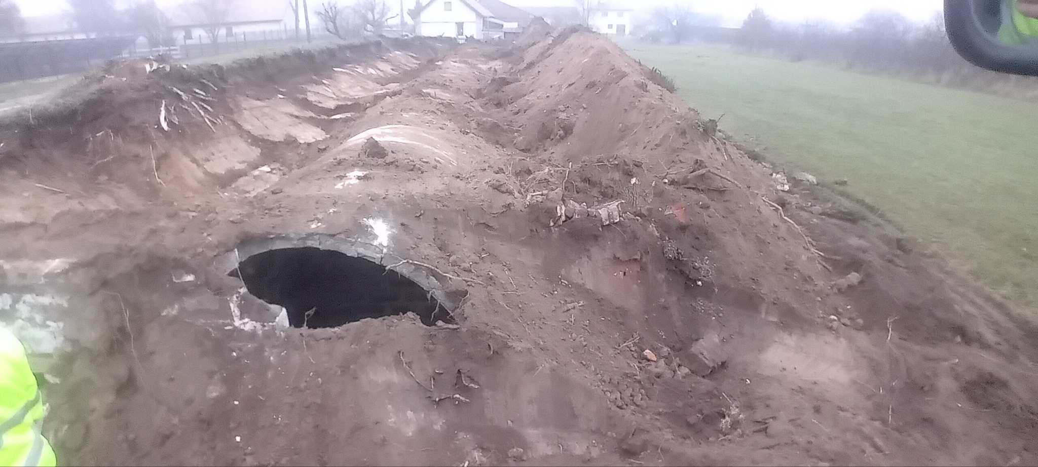 Wykonany wykop pod rurę przepustową, widoczne ślady po pracy koparki, ziemia usypana po bokach wykopu, w tle budynki mieszkalne i drzewa.