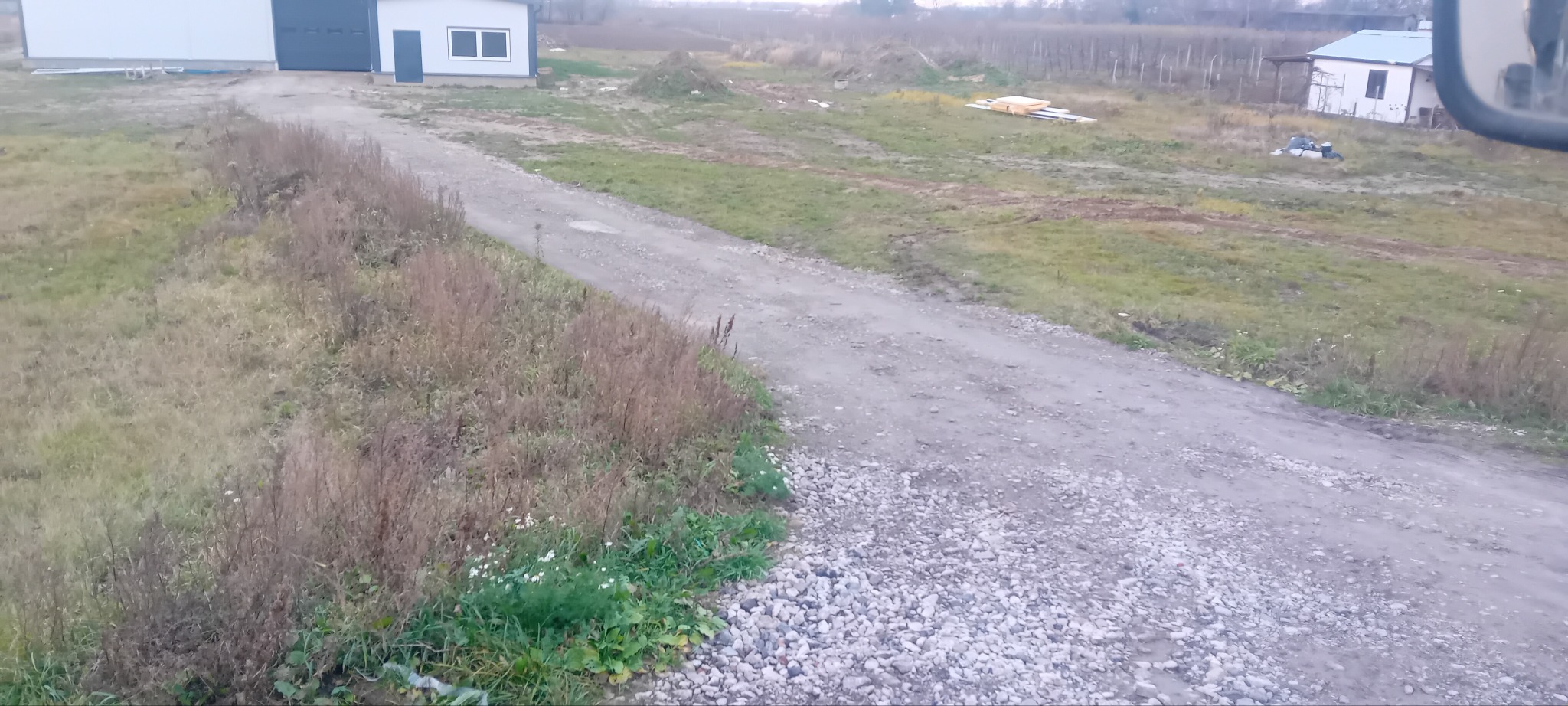 Utrwalona, szutrowa droga dojazdowa do budynku gospodarczego, otoczona polami i niską roślinnością. Widok z kabiny pojazdu.