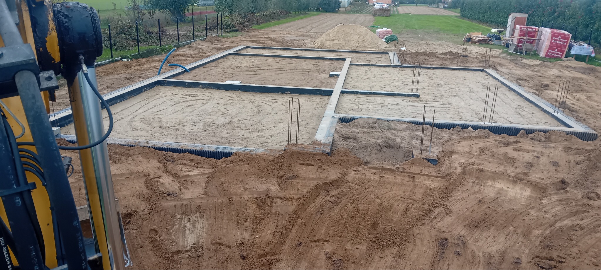 Fundamenty domu jednorodzinnego w trakcie budowy, widok z góry z perspektywy maszyny budowlanej. W tle ziemia, cegły i teren budowy.