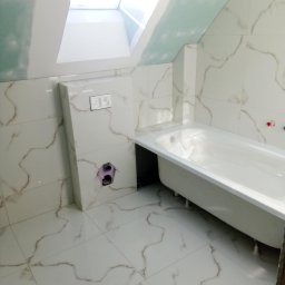 Remonty i Wykończenia Piotr Kliś - Biała wanna na nóżkach w wykończonej łazience na poddaszu, z oknem dachowym i marmurowymi płytkami. Widoczne przygotowane instalacje.