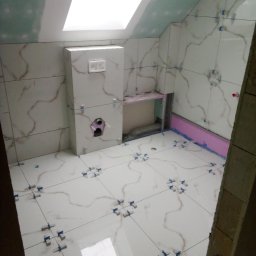 Remonty i Wykończenia Piotr Kliś - Białe płytki imitujące marmur na ścianach i podłodze łazienki na poddaszu w trakcie montażu. Widoczne krzyżyki dystansowe i fragment okna dachowego.