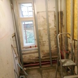 Remonty i Wykończenia Piotr Kliś - Remont pokoju: metalowy stelaż pod płyty gipsowe przy oknie, stara ściana z resztkami tapet, drabina malarska. Widoczne rury instalacji grzewczej.