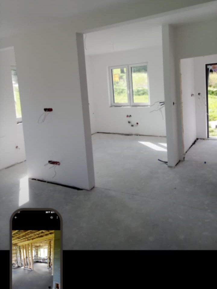 Surowe wnętrze z oknem i wystającymi kablami, przygotowane do dalszych prac wykończeniowych. W rogu ekran smartfona z widokiem na konstrukcję szkieletową.