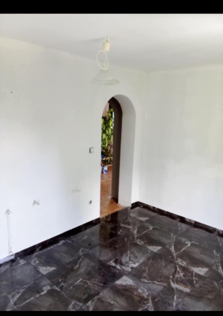 Białe ściany i ciemna marmurowa podłoga w pustym pokoju z łukowym przejściem do innego pomieszczenia. Widoczna lampa sufitowa i gniazdka elektryczne.