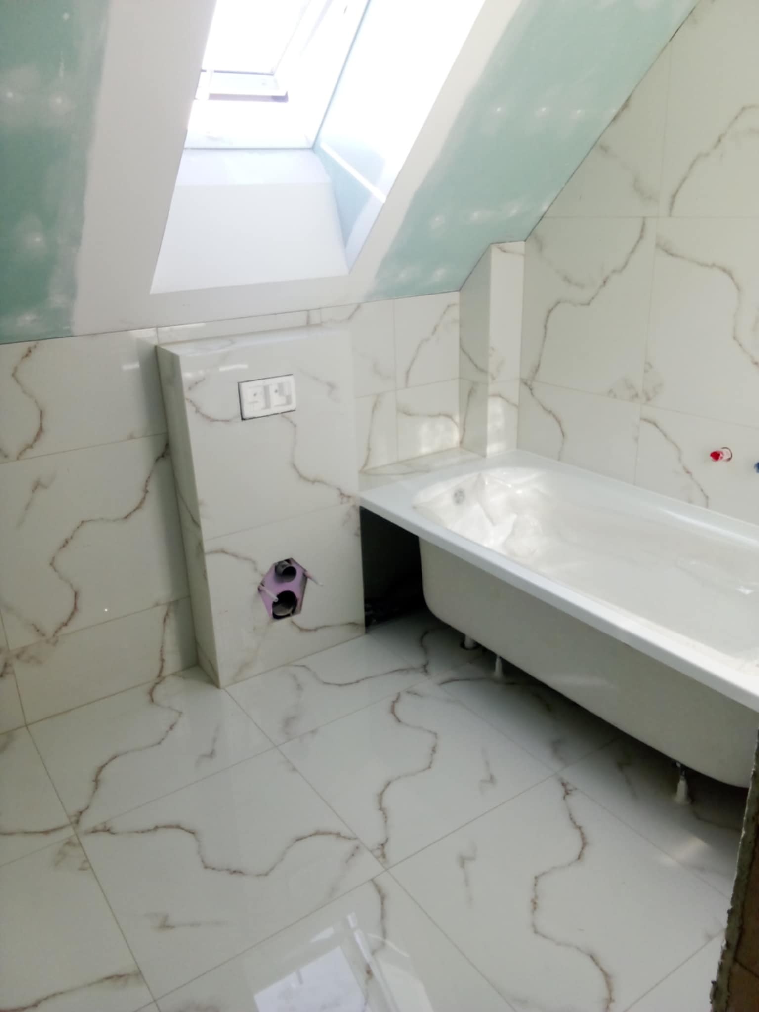 Biała wanna na nóżkach w wykończonej łazience na poddaszu, z oknem dachowym i marmurowymi płytkami. Widoczne przygotowane instalacje.