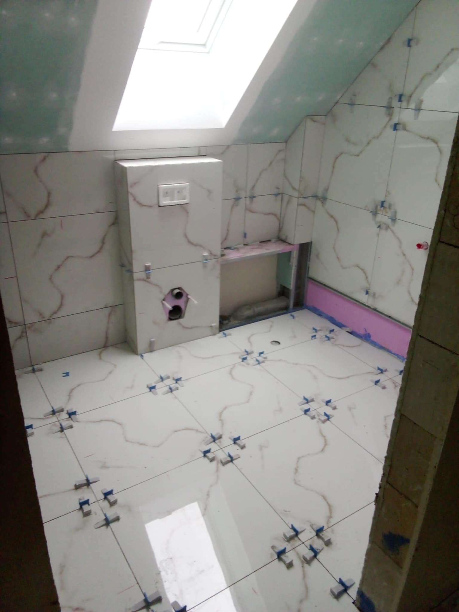 Białe płytki imitujące marmur na ścianach i podłodze łazienki na poddaszu w trakcie montażu. Widoczne krzyżyki dystansowe i fragment okna dachowego.