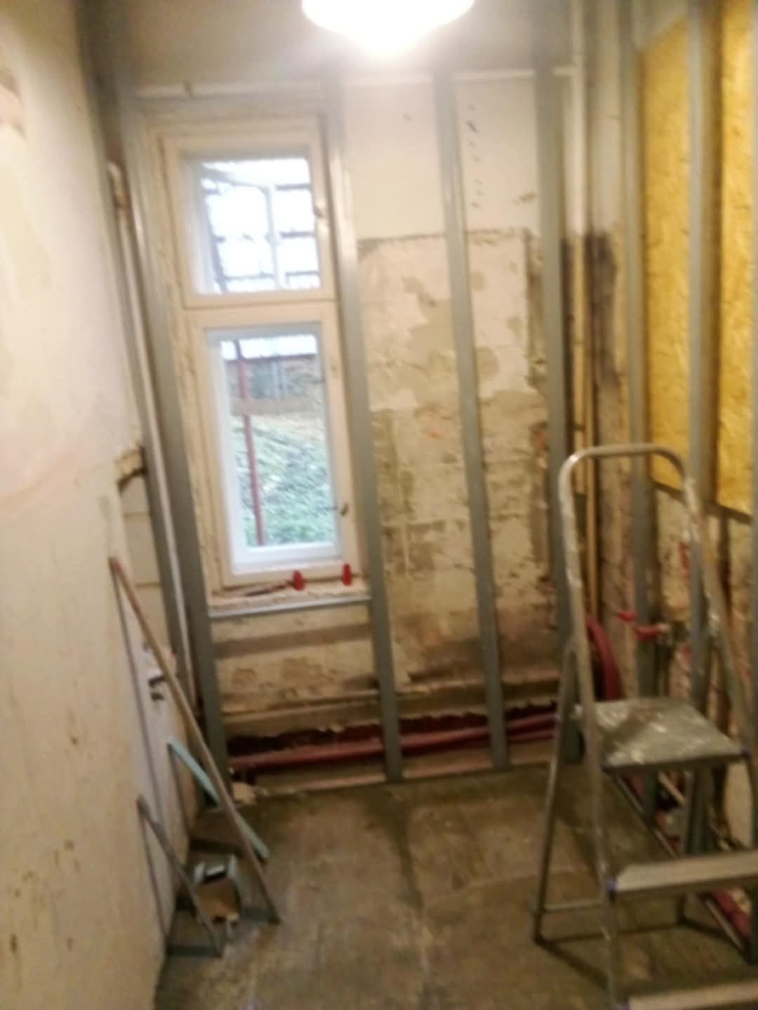 Remont pokoju: metalowy stelaż pod płyty gipsowe przy oknie, stara ściana z resztkami tapet, drabina malarska. Widoczne rury instalacji grzewczej.