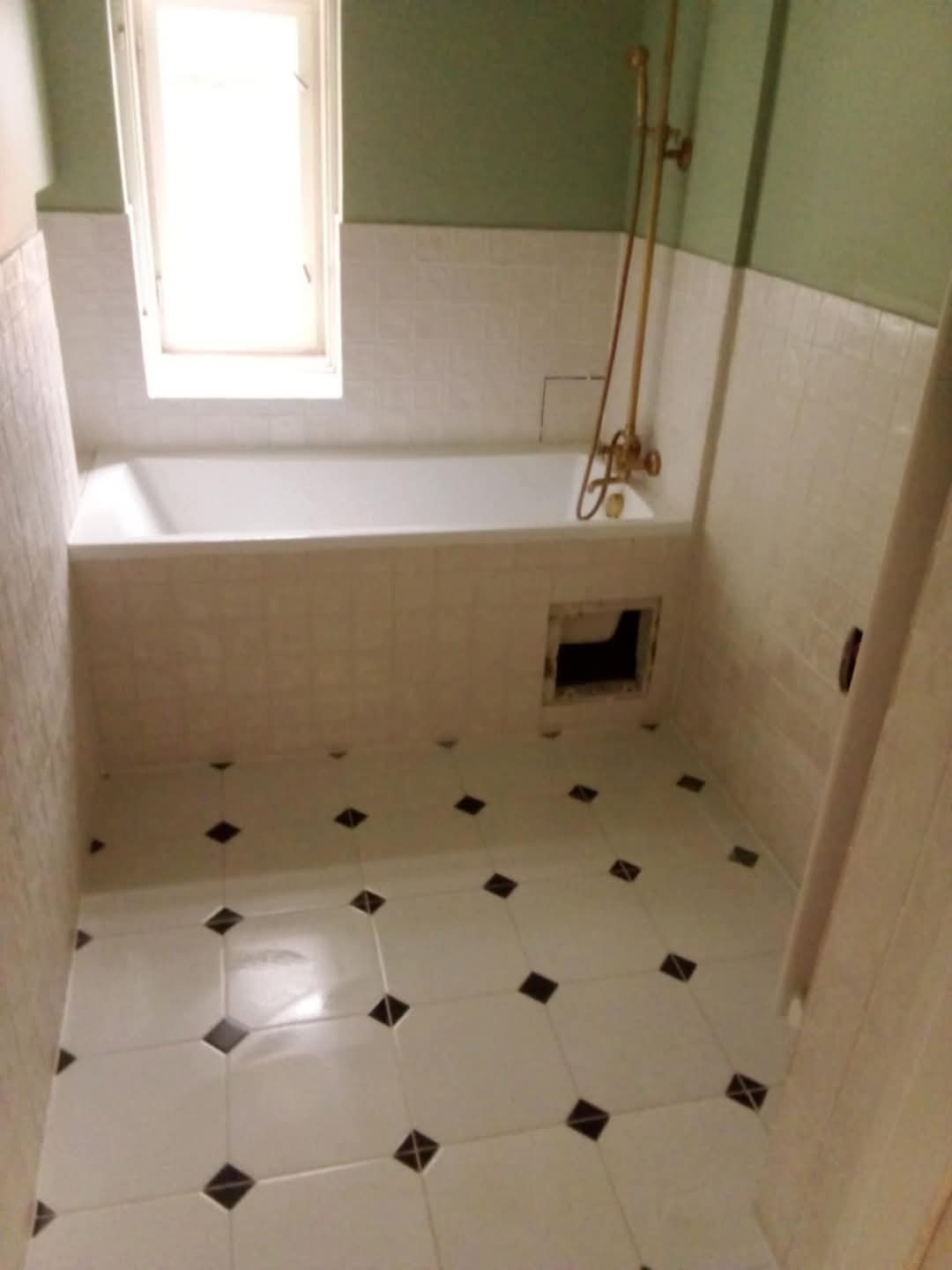 Wykończona łazienka z białą wanną obłożoną kafelkami, armaturą w kolorze złota i podłogą w biało-czarną szachownicę. Widoczny fragment ściany w odcieniach zieleni.