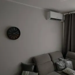 Biały klimatyzator ścienny zamontowany w szarym pokoju nad szarą kanapą, obok ciemny zegar ścienny z drewnianymi wskazówkami.