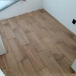 Ułożone podłużne panele podłogowe imitujące drewno, w wąskim pomieszczeniu z białymi ścianami i czarnym gniazdkiem elektrycznym.