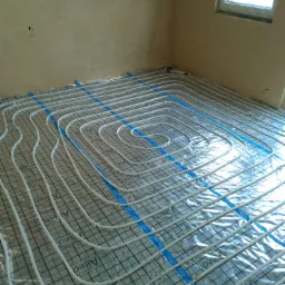 Białe rury ogrzewania podłogowego ułożone w pętle na folii z nadrukiem siatki, przymocowane niebieską taśmą do podłoża w pomieszczeniu z oknem.