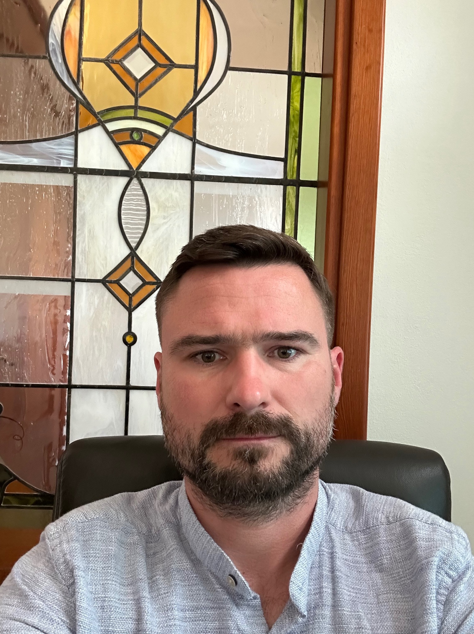Portret mężczyzny z brodą na krześle, na tle okna ze szkła witrażowego z geometrycznymi wzorami w odcieniach brązu, beżu i zieleni.
