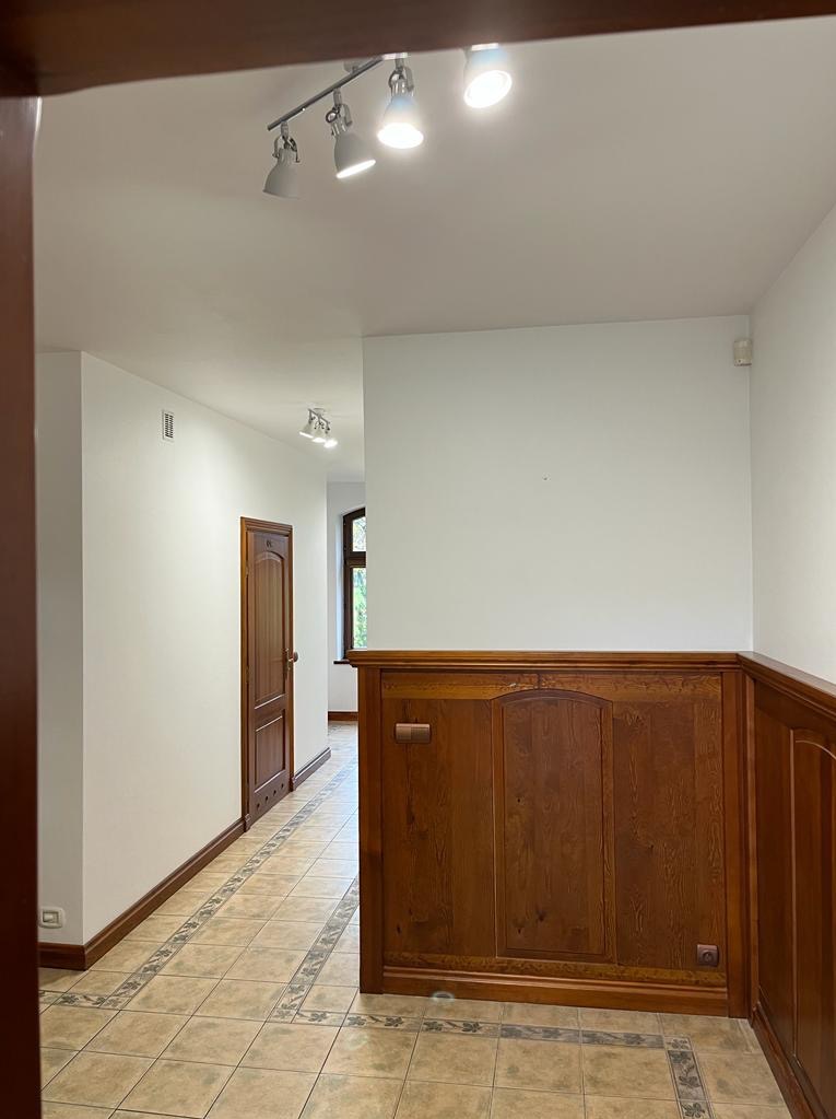 Korytarz z drewnianą boazerią i płytkami podłogowymi w tonacji beżu. Widoczne drzwi i lampy sufitowe. W tle okno z widokiem na zieleń.